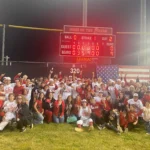 A group of excited baseball players celebrating a victory on the field at night, wearing white t-shirts with 'Champions' printed on them.